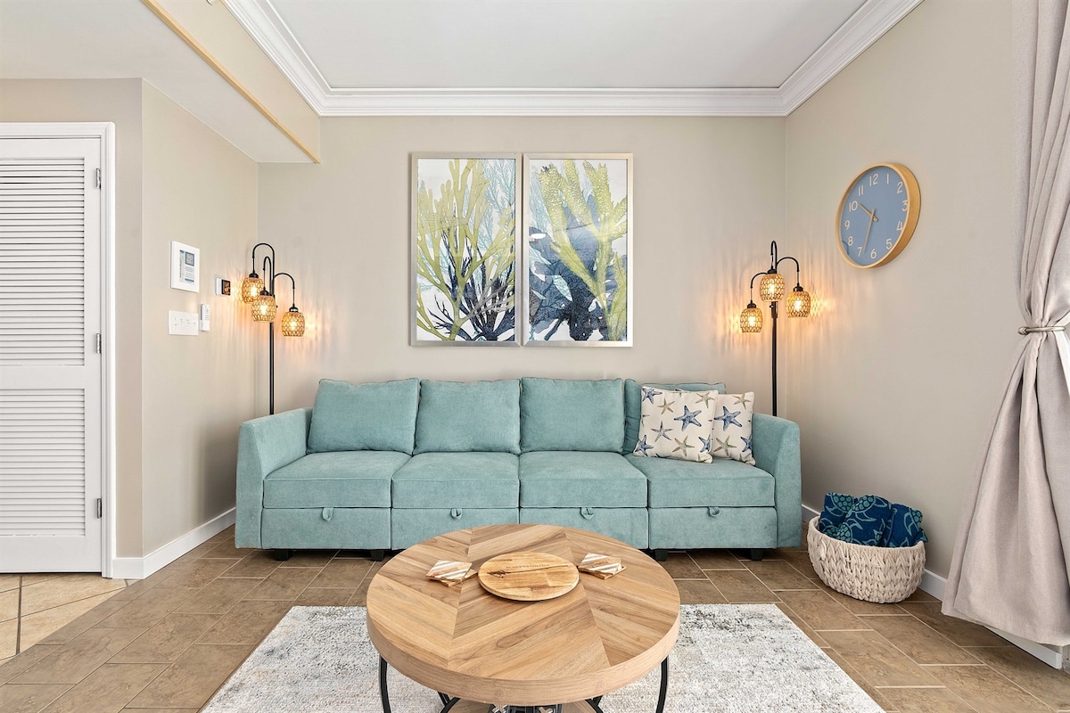 A comfortable seating area features a soft, light blue sectional sofa adorned with decorative pillows. A round wooden coffee table sits in the center, accompanied by a woven basket. Two framed botanical prints are displayed on the wall, with wall sconces providing gentle illumination.