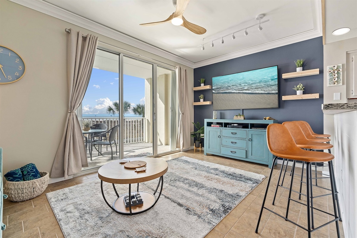 A bright living area features a turquoise media console with a large screen showcasing coastal imagery. Two bar stools offer additional seating at the kitchen counter. Light flows in through sliding glass doors, leading to a balcony with a view of palm trees.