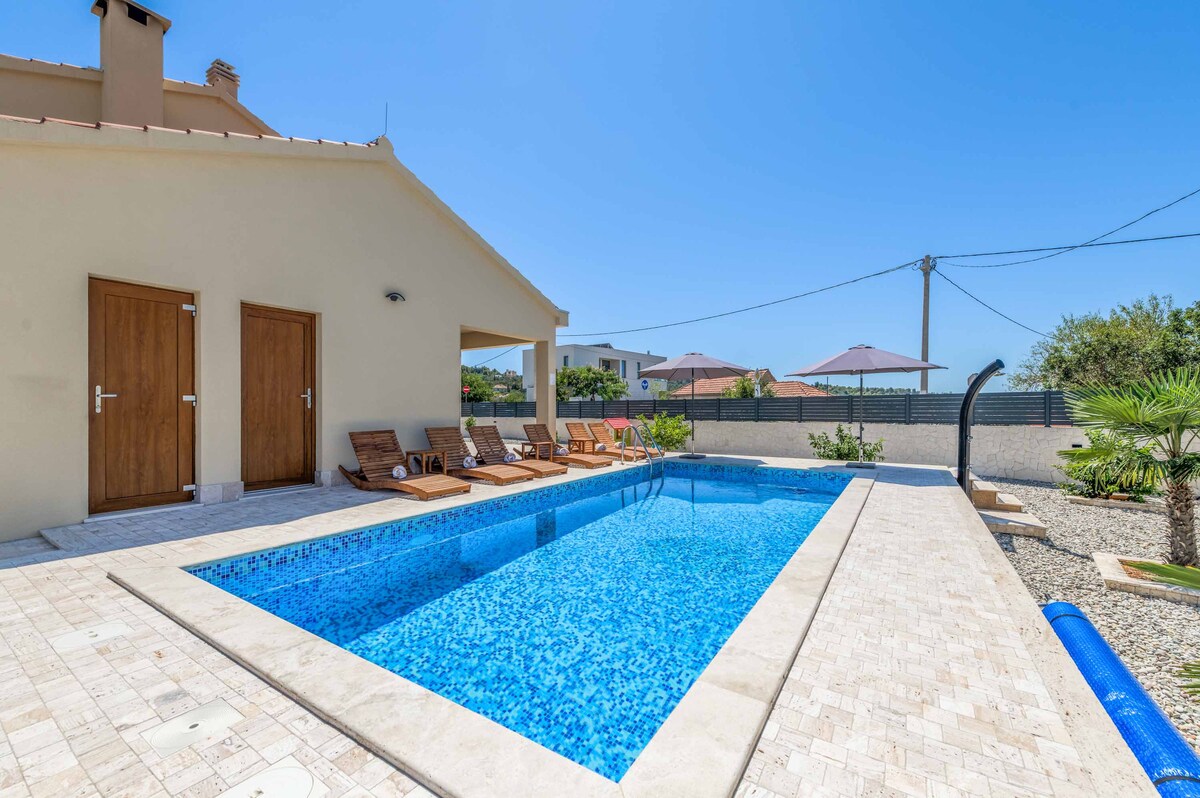 A serene outdoor space features a sparkling swimming pool surrounded by light-colored stone tiles. Sun loungers are positioned along one side, inviting relaxation. Two wooden doors lead to the interior, while lush greenery and shaded umbrellas provide a refreshing backdrop.