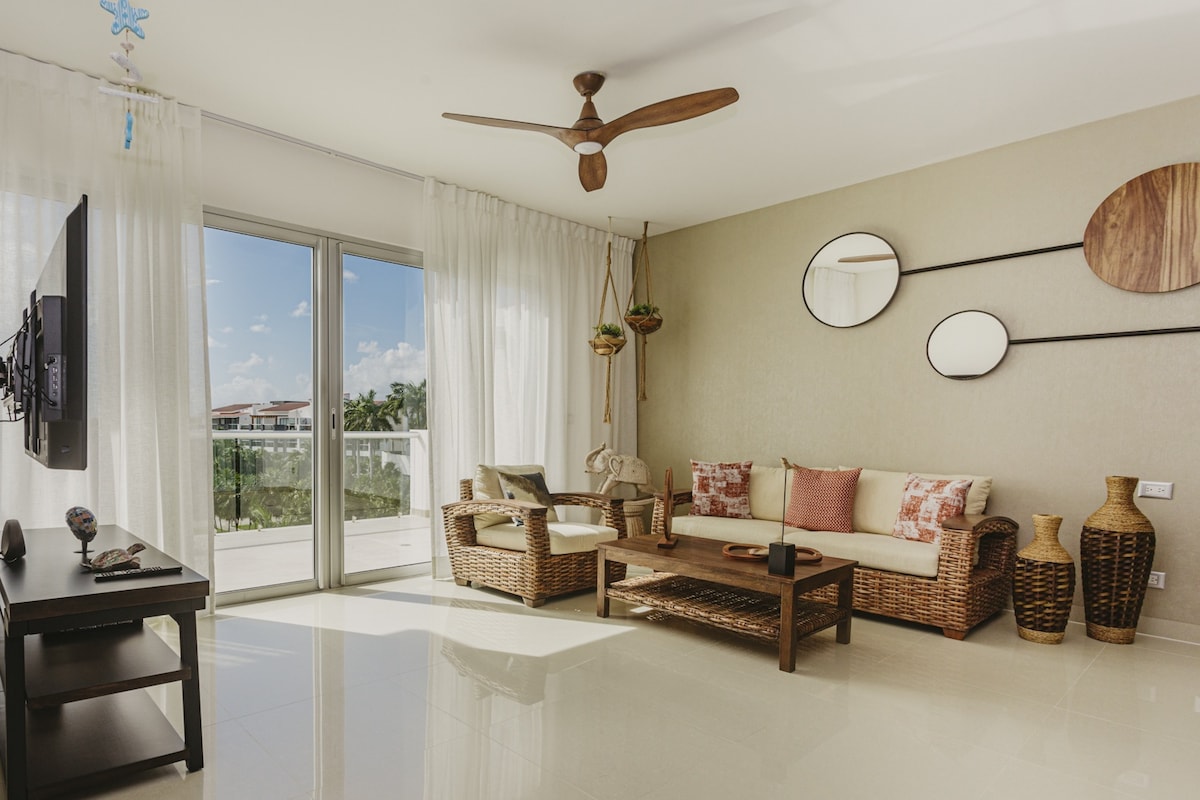 A living area features a comfortable seating arrangement with wicker furniture and decorative cushions. A television is mounted on the wall, and natural light filters through sheer curtains. A decorative wall arrangement includes circular mirrors and wooden elements, enhancing the overall space.