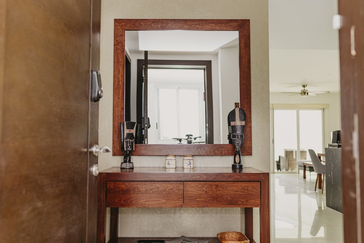 A wooden console table is positioned beneath a large mirror, enhancing the entryway's sense of space. Decorative items flank the mirror, while the soft, neutral tones of the walls create a welcoming atmosphere. Natural light is visible from the adjoining living area.