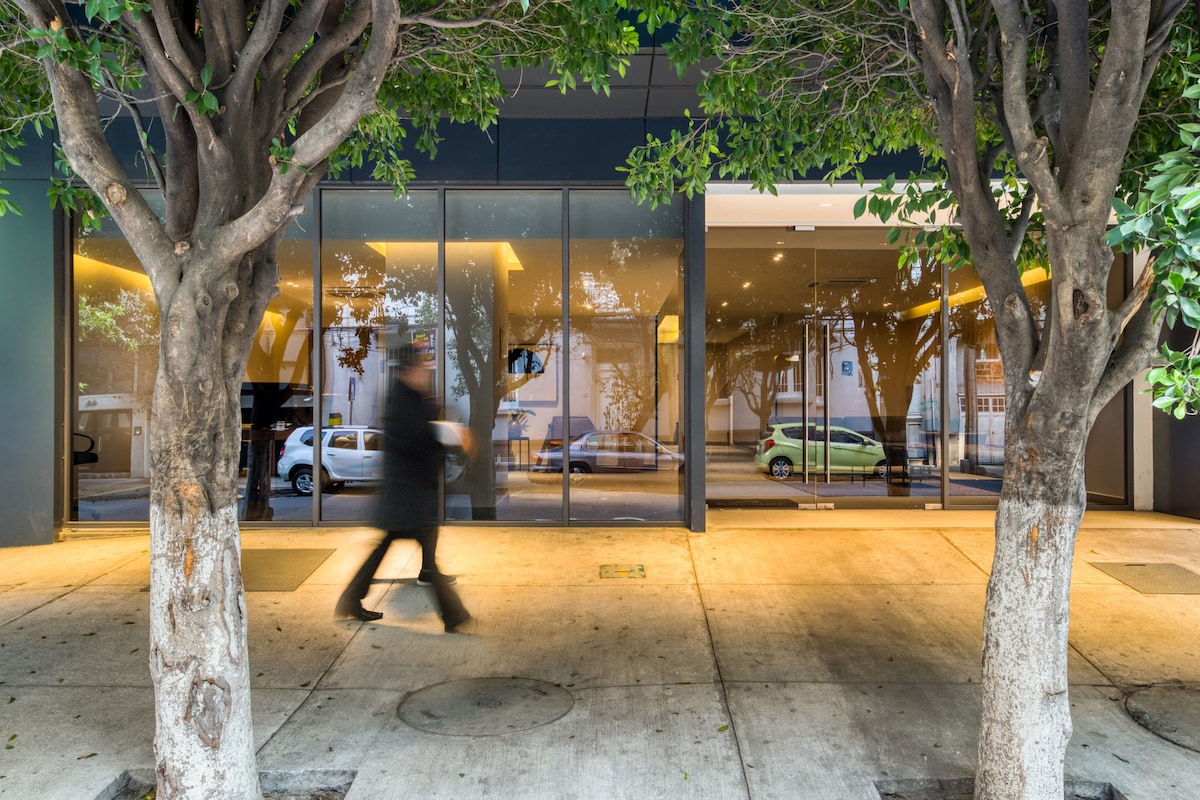 A modern building entrance is framed by leafy trees on either side. Large glass windows provide a glimpse of the interior space, reflecting the surrounding street activity. Sidewalks are visible, with a blurred figure walking past, suggesting a vibrant urban atmosphere.