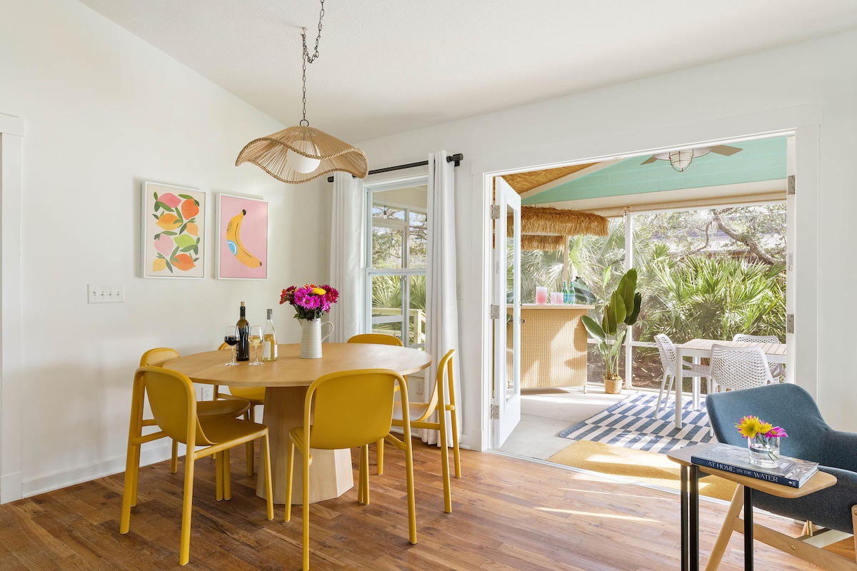 A bright dining area is furnished with a round wooden table surrounded by four yellow chairs. Colorful artwork adorns the walls, while large glass doors open to a screened porch featuring outdoor seating and tropical plants. Natural light enhances the cheerful ambiance.