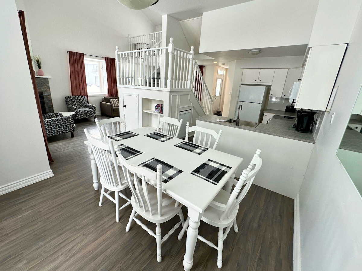 A bright dining area features a large white table surrounded by eight chairs, designed for group meals. The space is complemented by a cozy sitting area with patterned armchairs and a staircase leading to an upper level, enhancing the open layout.