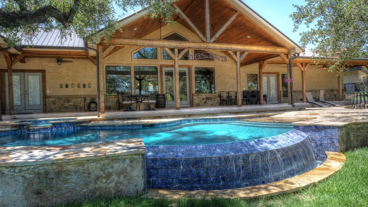 An outdoor view of the home showcases a spacious infinity pool with blue tiles, flowing water, and a sunlit patio area. The structure features a wooden overhang, highlighting large glass doors and a stone facade, creating a well-integrated indoor and outdoor atmosphere.