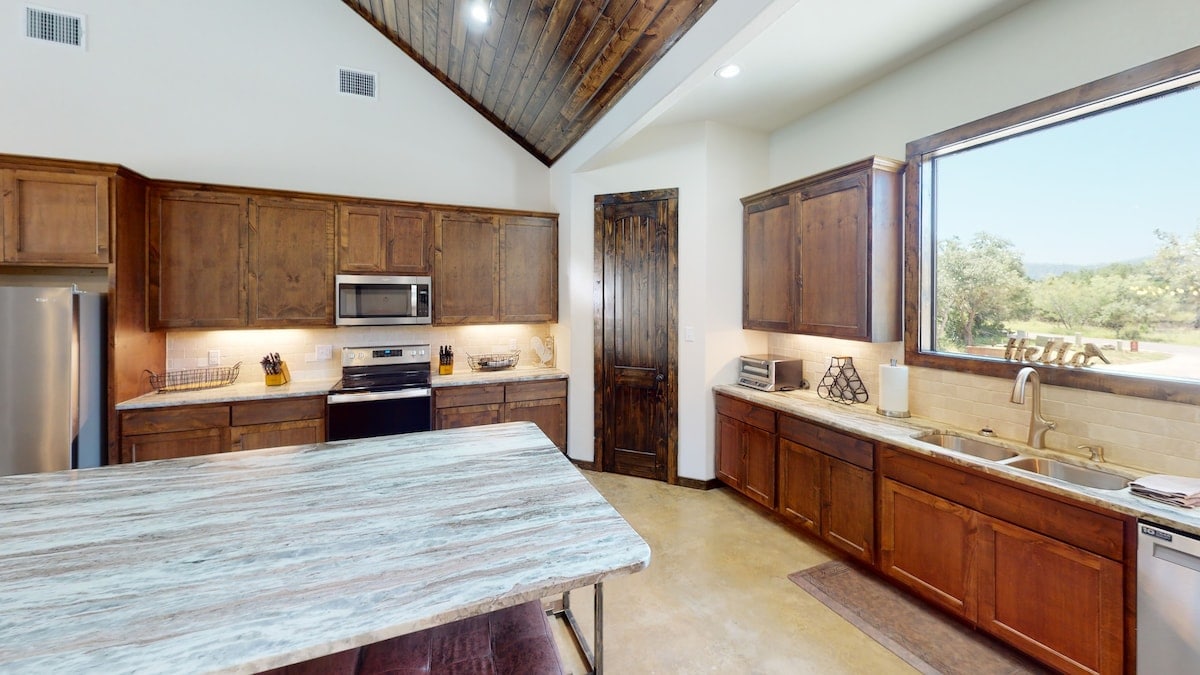 A spacious kitchen features wooden cabinetry and stainless steel appliances. A large island with a textured countertop provides additional workspace. Natural light fills the area through a large window, offering views of the outdoor scenery. The ceiling showcases wood beams for added character.