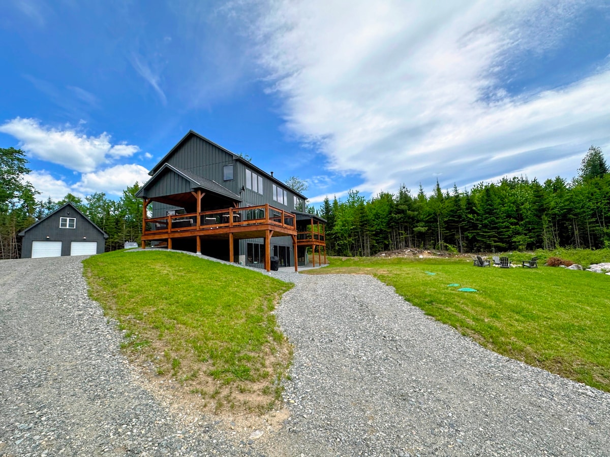 A modern rustic home is showcased with spacious decks and large windows, surrounded by green lawns and towering trees. A gravel pathway leads up to the house, while a cozy seating area is visible in the backyard under a clear blue sky.