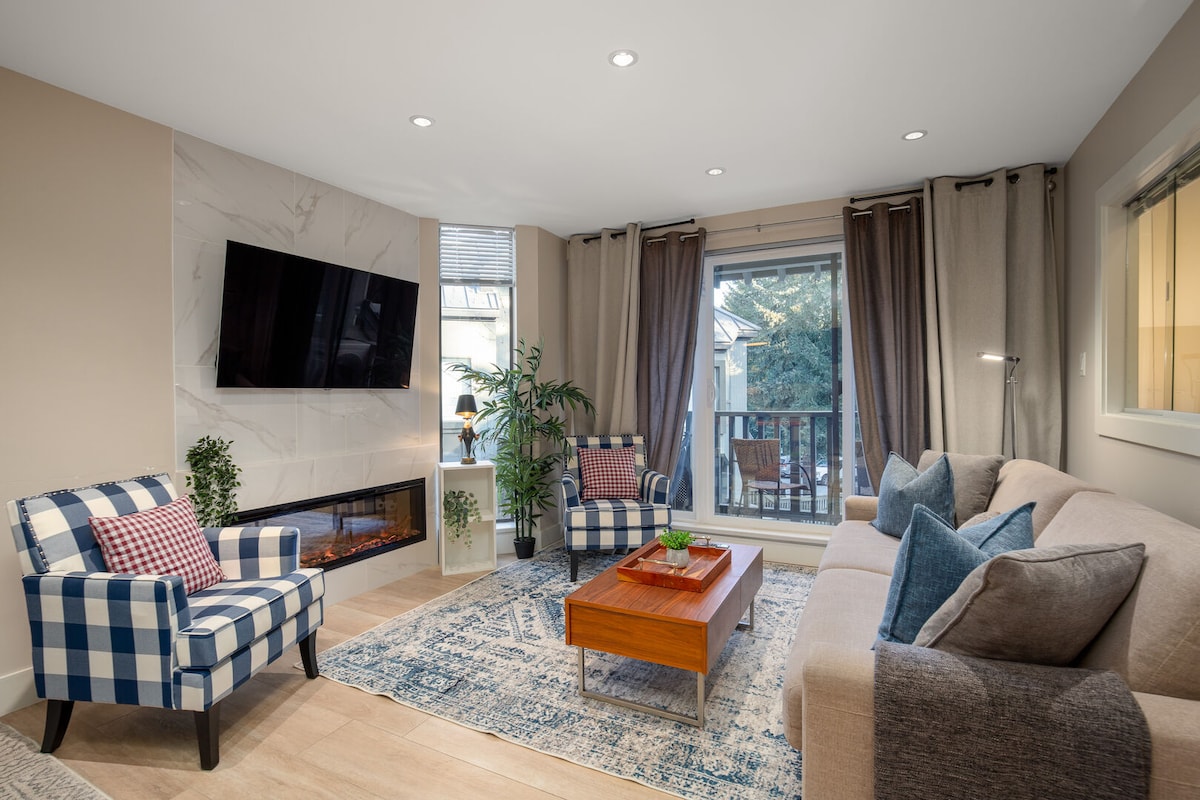 The living area features a smart TV mounted on a light-colored wall, with a modern electric fireplace below. A warm-toned wooden coffee table is centered on a decorative area rug. Two checkered armchairs provide seating, and a large window offers views of the private balcony.
