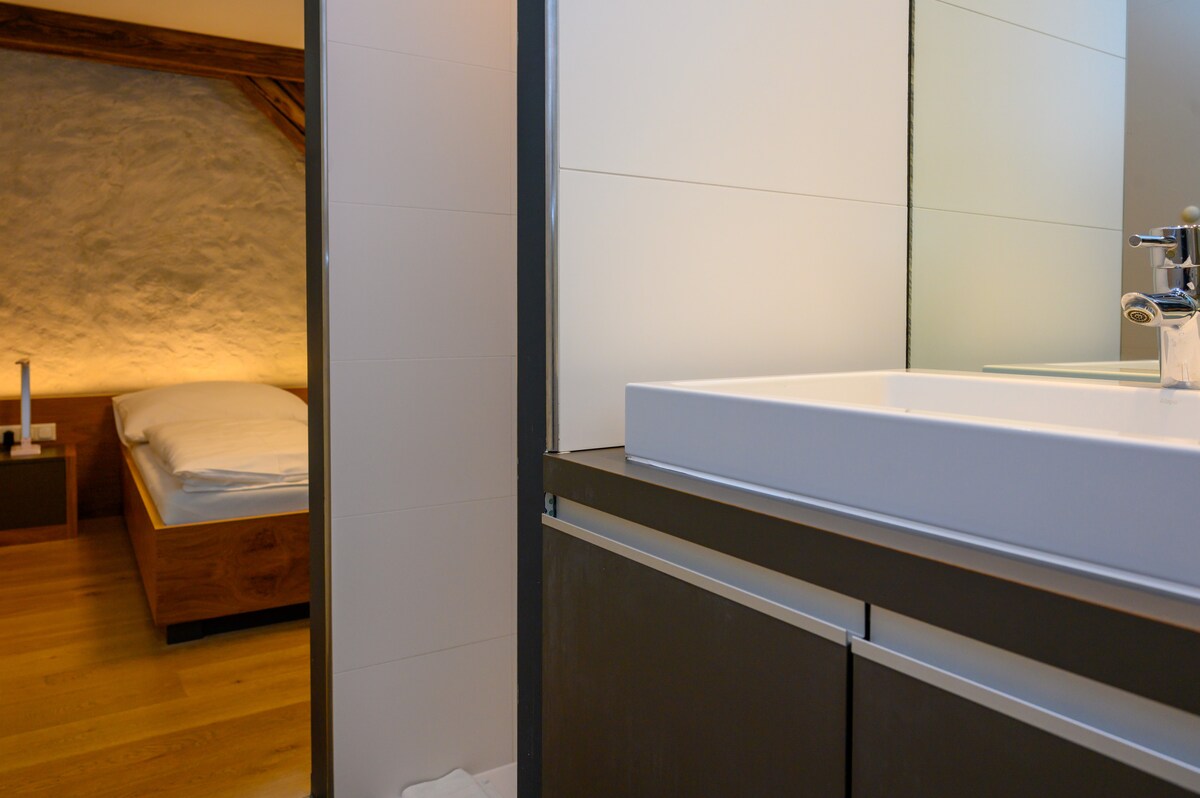 A modern bathroom features a sleek, white sink atop dark cabinetry. Light reflects off the walls, leading into a cozy sleeping area with natural wood accents and a neatly made bed, subtly illuminated by warm lighting.