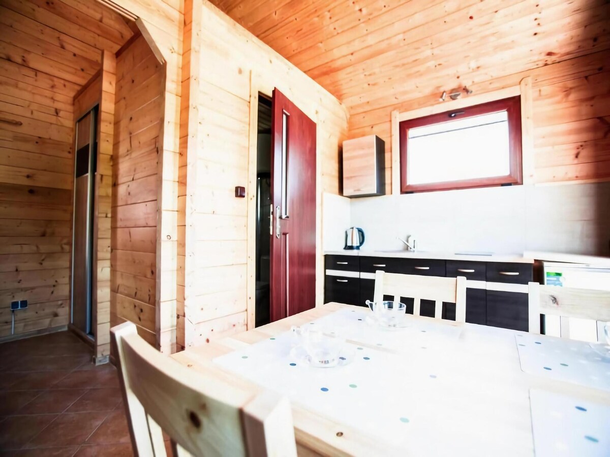 A warm wooden interior features a dining area with a table set for four. The open kitchen includes essential appliances, and large windows allow natural light to fill the space, illuminating the rich wood walls and providing a cozy ambiance.