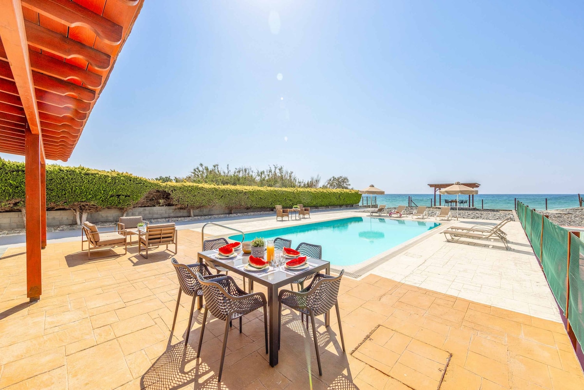 An outdoor area surrounds a private swimming pool, complemented by a spacious terrace. A dining table is set with dishes, and sun loungers are arranged along the poolside, providing relaxation options. Palms and greenery frame the scene against a clear blue sky and distant ocean.