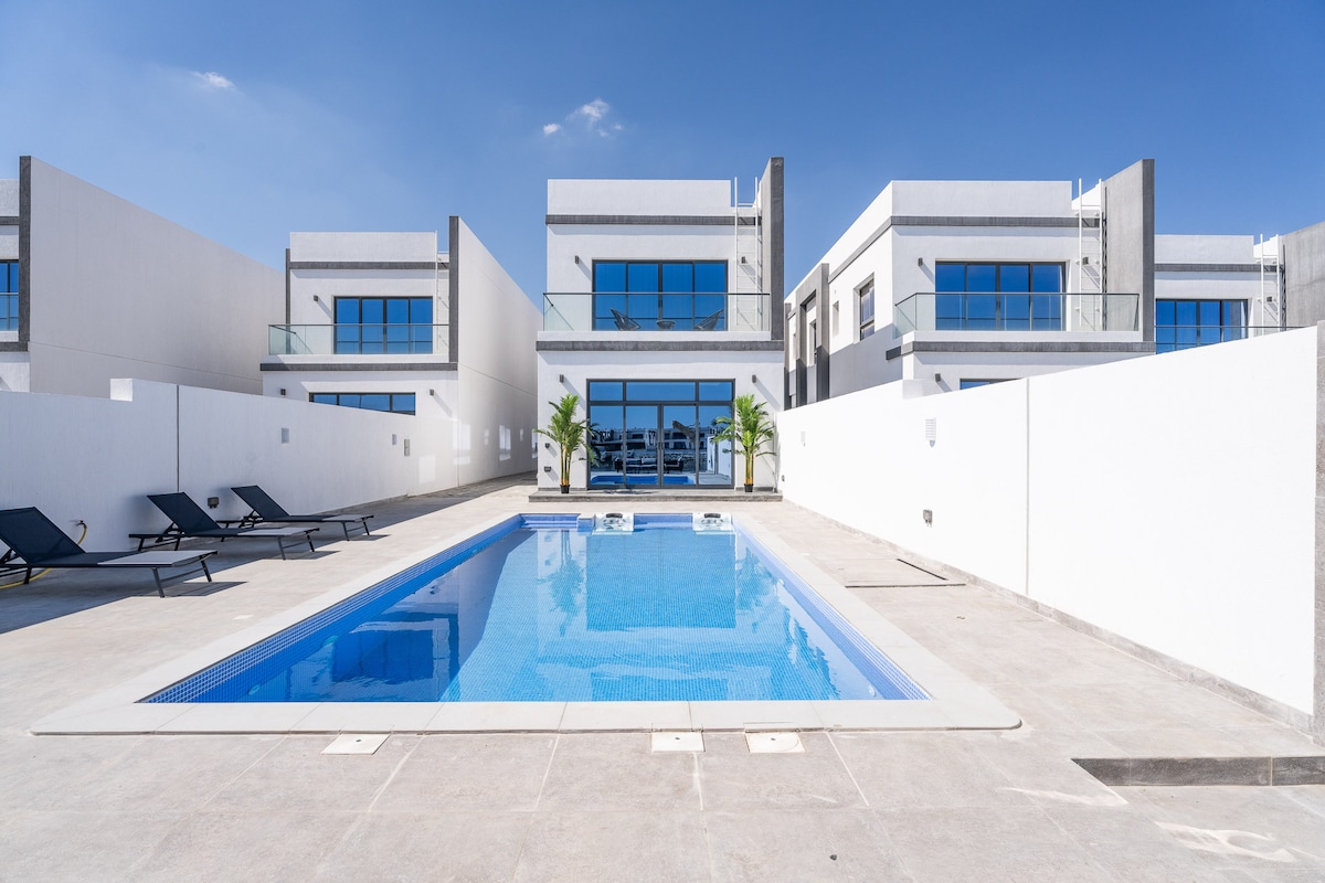 A modern villa is showcased, featuring a sparkling pool surrounded by lounge chairs. The exterior design highlights large glass windows that offer views into the interior, with white walls creating a clean and spacious outdoor area.