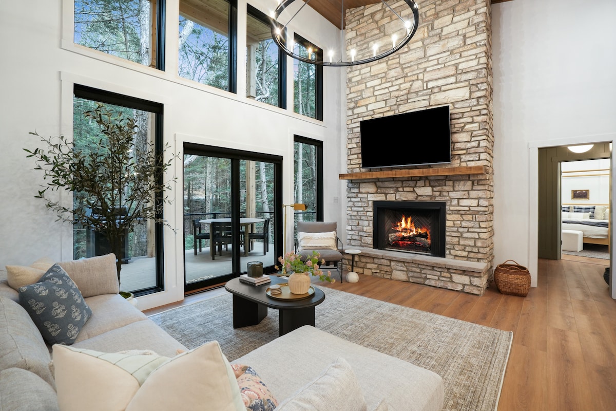 A modern living area features a comfortable couch with decorative pillows, positioned in front of a stone fireplace. Large windows invite natural light and showcase the outdoor scenery. A flat-screen TV is mounted above the fireplace, and a small table is adorned with decorative items.