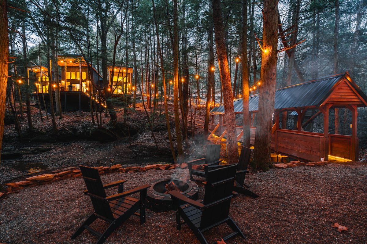A designated outdoor area features a circular fire pit surrounded by several black wooden chairs. Twinkling lights are strung between the trees, illuminating the pathway leading to a cabin in the background. Lush greenery complements the serene setting.