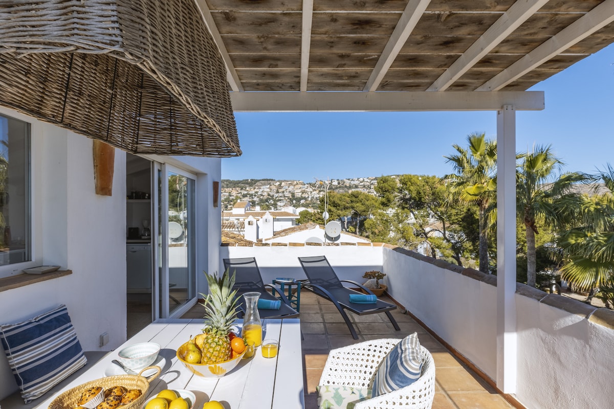 An outdoor terrace is showcased, featuring a dining table adorned with fruits and beverages. Two sunbeds are positioned nearby under a wooden pergola, with views extending to palm trees and distant hills. Bright sunlight accentuates the inviting space.