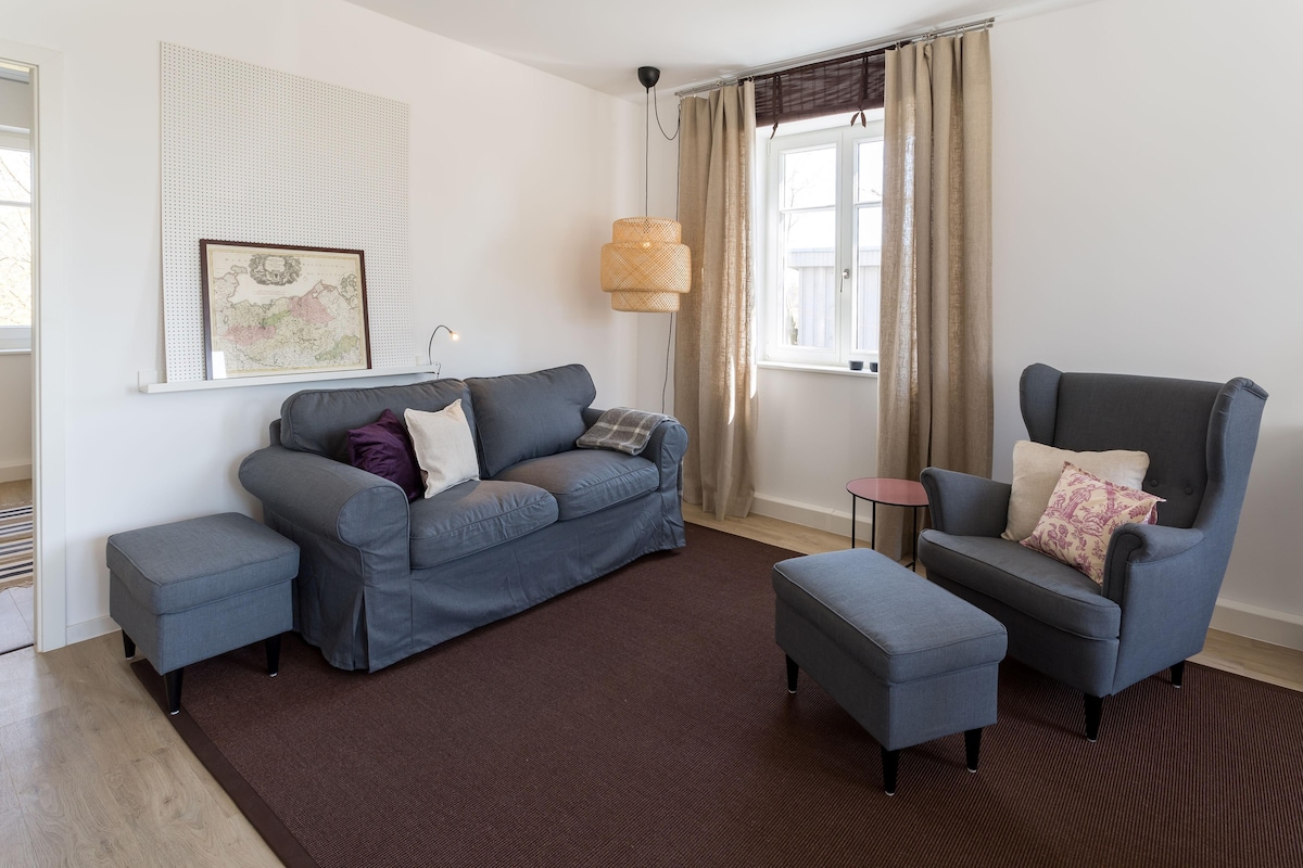 A spacious living area features a comfortable blue sofa adorned with decorative cushions. A matching ottoman complements a wingback chair. Natural light enters from a nearby window, surrounded by light-colored curtains, enhancing the welcoming atmosphere. A small side table is positioned next to the chair.
