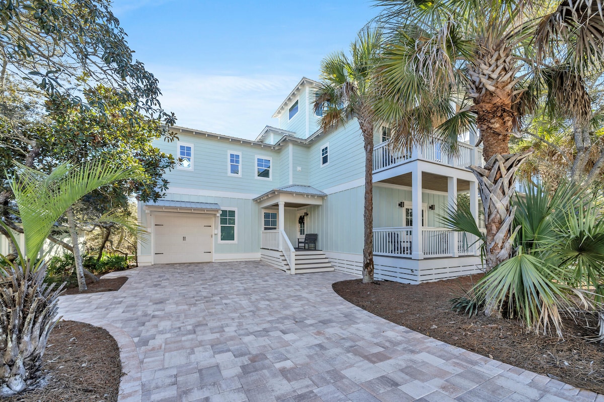 The exterior of the home is highlighted by light blue siding and brick-paver driveway. Tropical landscaping features palm trees and dense greenery. A spacious front porch with seating invites relaxation. A one-car garage is visible on the left side, enhancing the home's curb appeal.