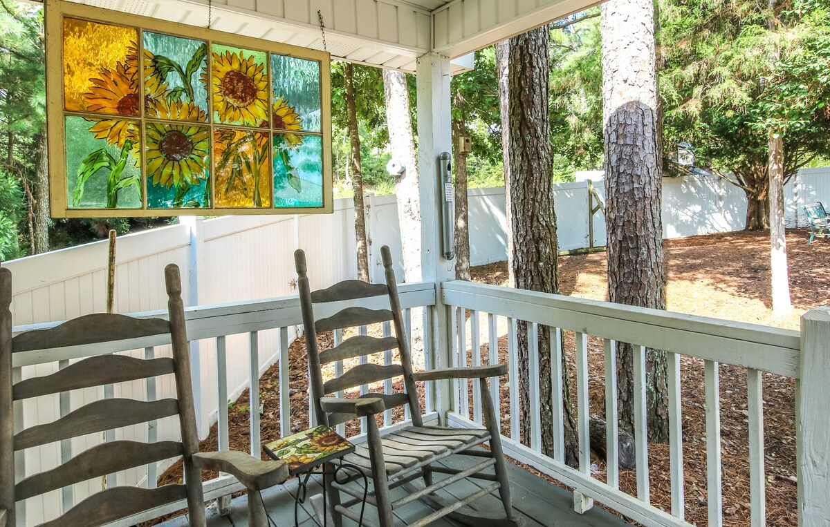 A serene screened porch features two wooden rocking chairs, positioned to offer views of the surrounding greenery. A stained glass art piece depicting sunflowers adds a splash of color, while the natural light filters through, creating a peaceful ambiance.