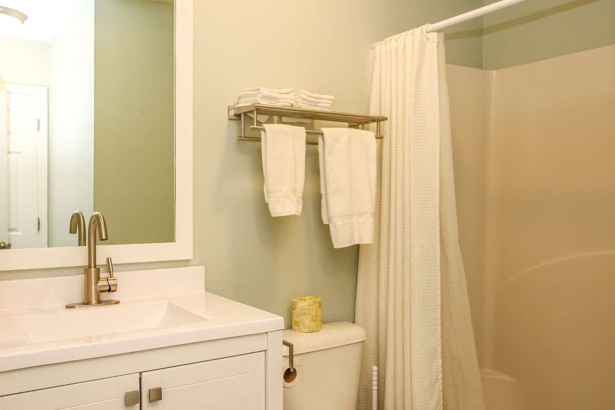 The bathroom features a light green wall, a modern sink with a sleek faucet, and a large mirror. Fresh white towels hang neatly on a rack, while a cream-colored shower curtain complements the neutral tones. A small decorative item is placed atop the toilet.