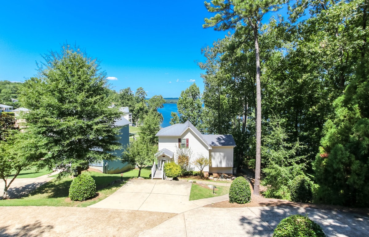The exterior of the house is framed by lush greenery and features a light-colored façade. A paved driveway leads to the entrance, while scenic lake views and blue skies are visible in the background, enhancing the peaceful setting surrounded by nature.