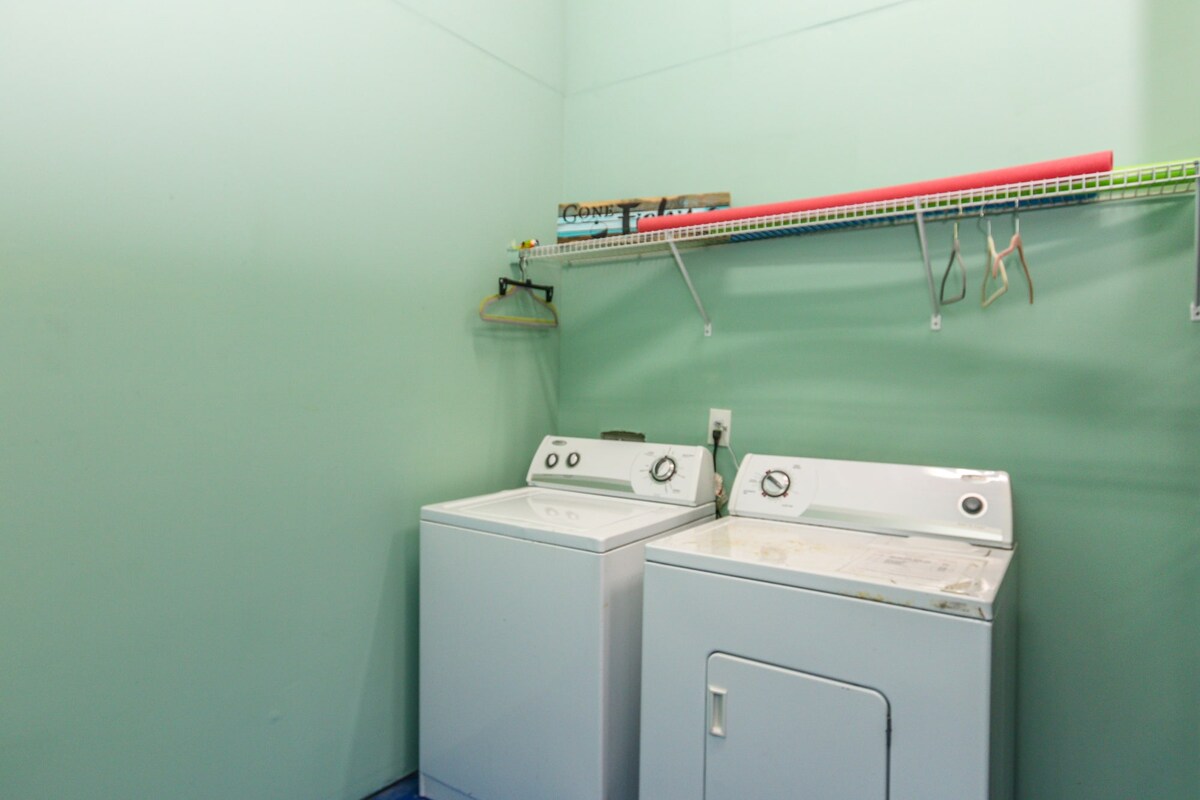 A laundry room features two white appliances: a washing machine and a dryer. The walls are painted a soft green, and a shelf above holds various items, including hangers and a rolled mat. The clean space offers practical amenities for guests.