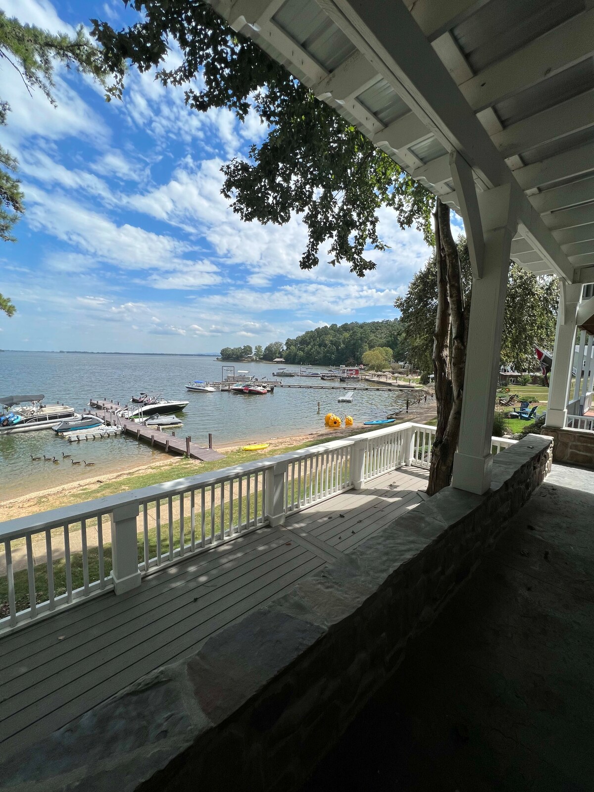 A scenic view of the lake is displayed from the porch area, featuring a sandy beach and boats docked nearby. The shoreline is lined with trees and grass, creating a natural setting. Sunny skies and fluffy clouds enhance the tranquil atmosphere.