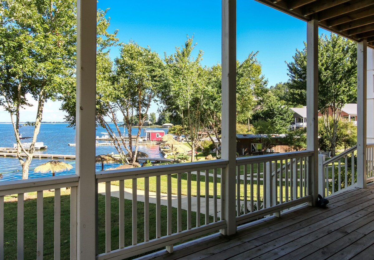 A covered porch is visible, adorned with railing that offers a view of the lake and surroundings. The serene water sparkles under sunlight, framed by trees and a grassy area, while a private dock and boats can be seen in the distance.
