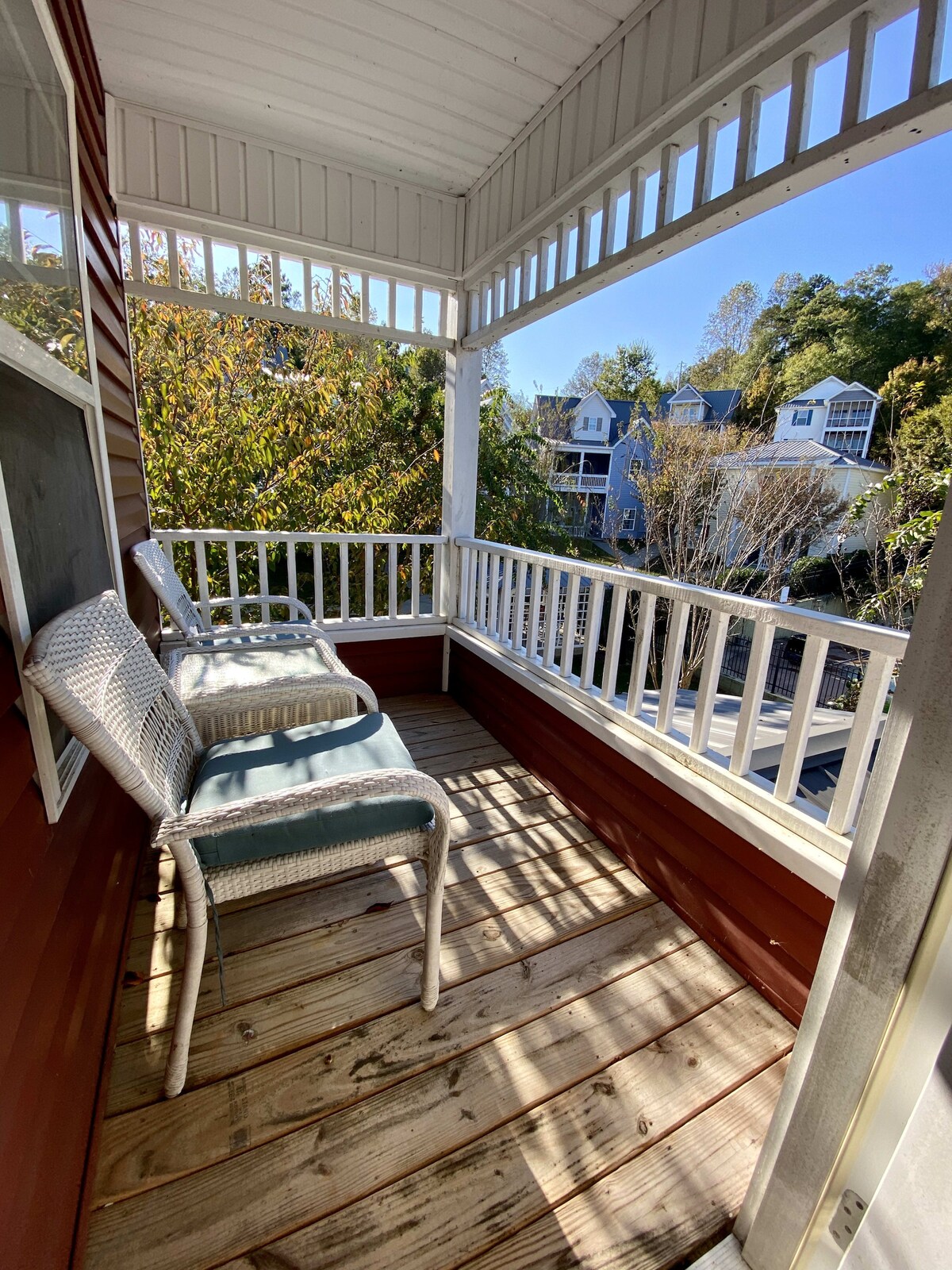 A cozy balcony features two white chairs, offering a space to relax. The wooden floor is illuminated by sunlight, and views of surrounding trees and neighboring houses can be seen through the railings. The setting provides a peaceful outdoor area.
