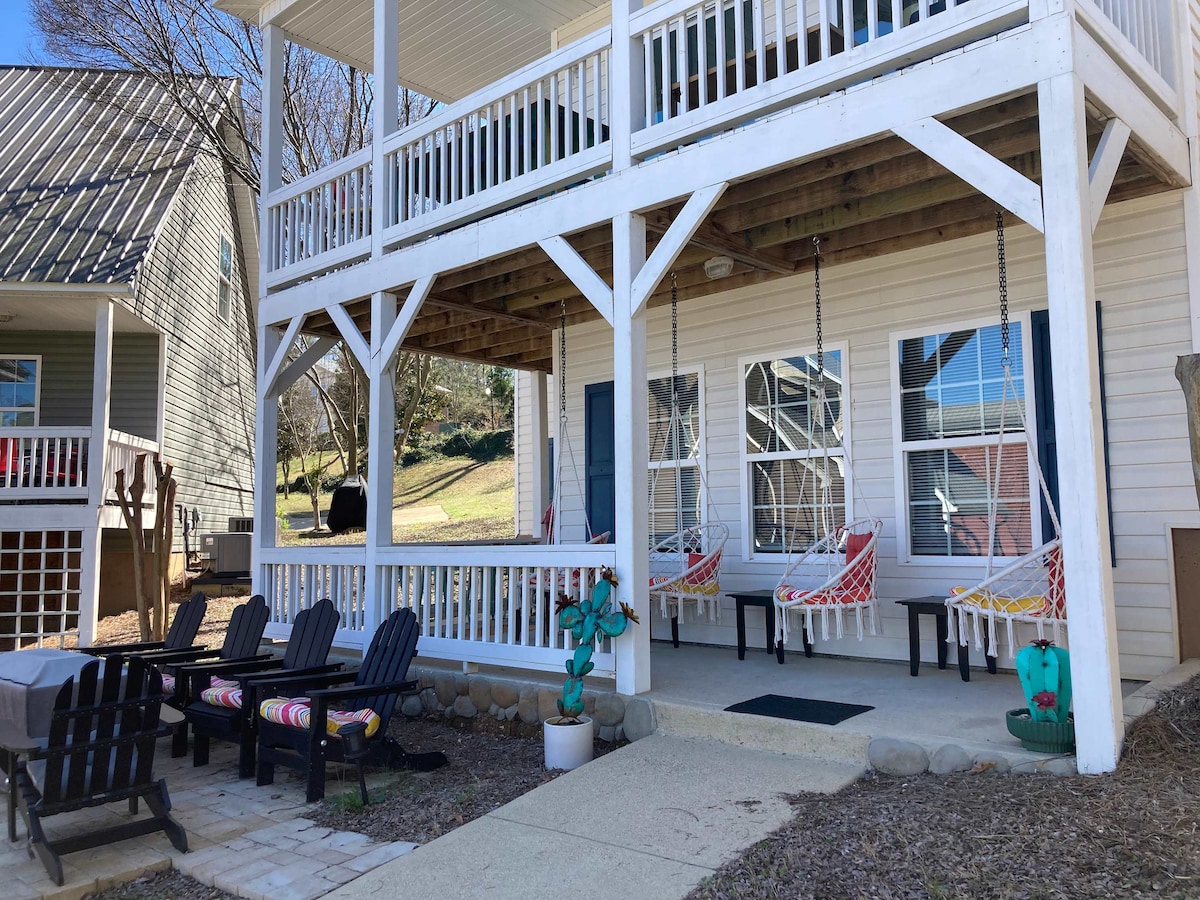 A welcoming outdoor space features a covered porch with hanging swings and seating arrangements. A seating area of black Adirondack chairs is positioned on the patio, complemented by a small table, inviting relaxation. Sunlight filters through the trees, creating a serene ambiance.