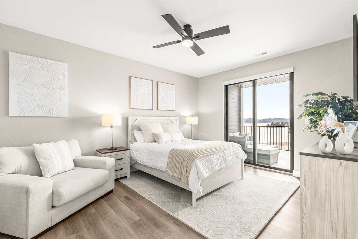 A serene bedroom features a spacious bed with light bedding and soft textures, complemented by a comfortable lounge chair. Large sliding doors lead to a private balcony, while subtle wall artwork adds to the calming ambiance.