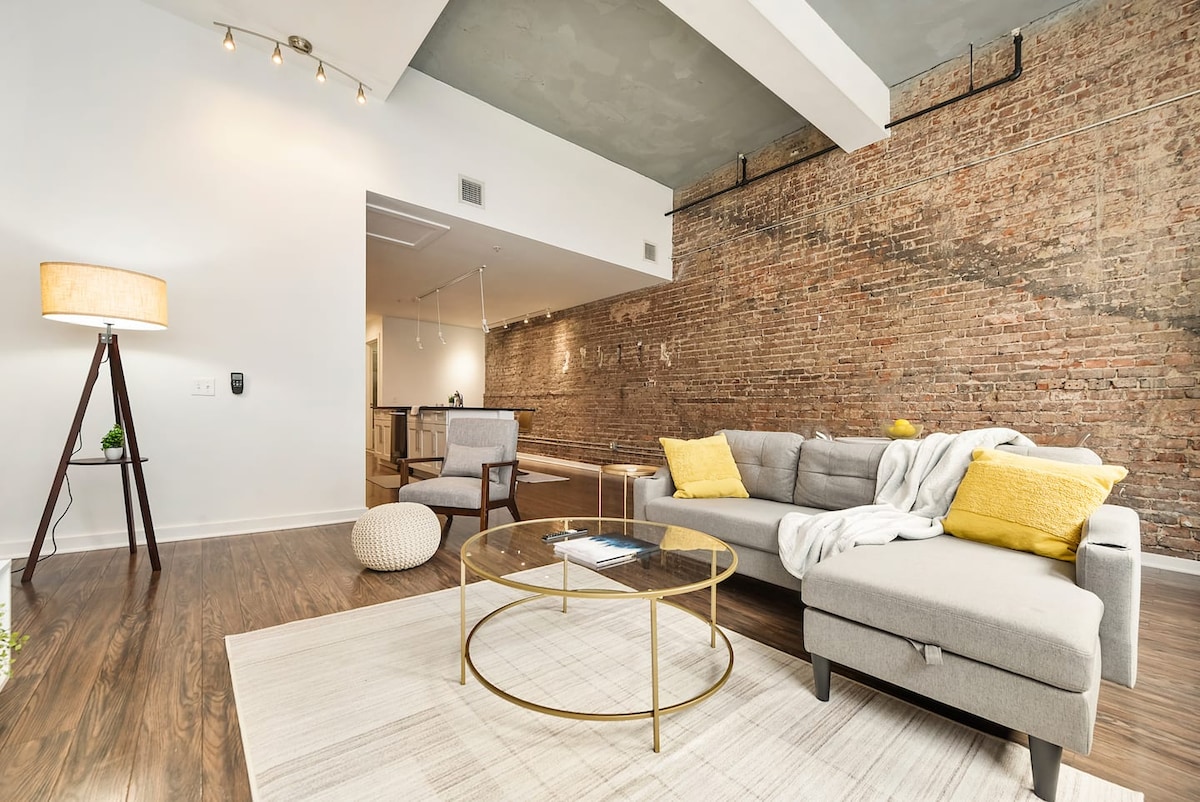 The living area features a modern grey sectional sofa adorned with yellow accent pillows. A round coffee table sits in front, topped with a magazine. Exposed brick walls add texture, while soft lighting from a floor lamp enhances the cozy atmosphere.