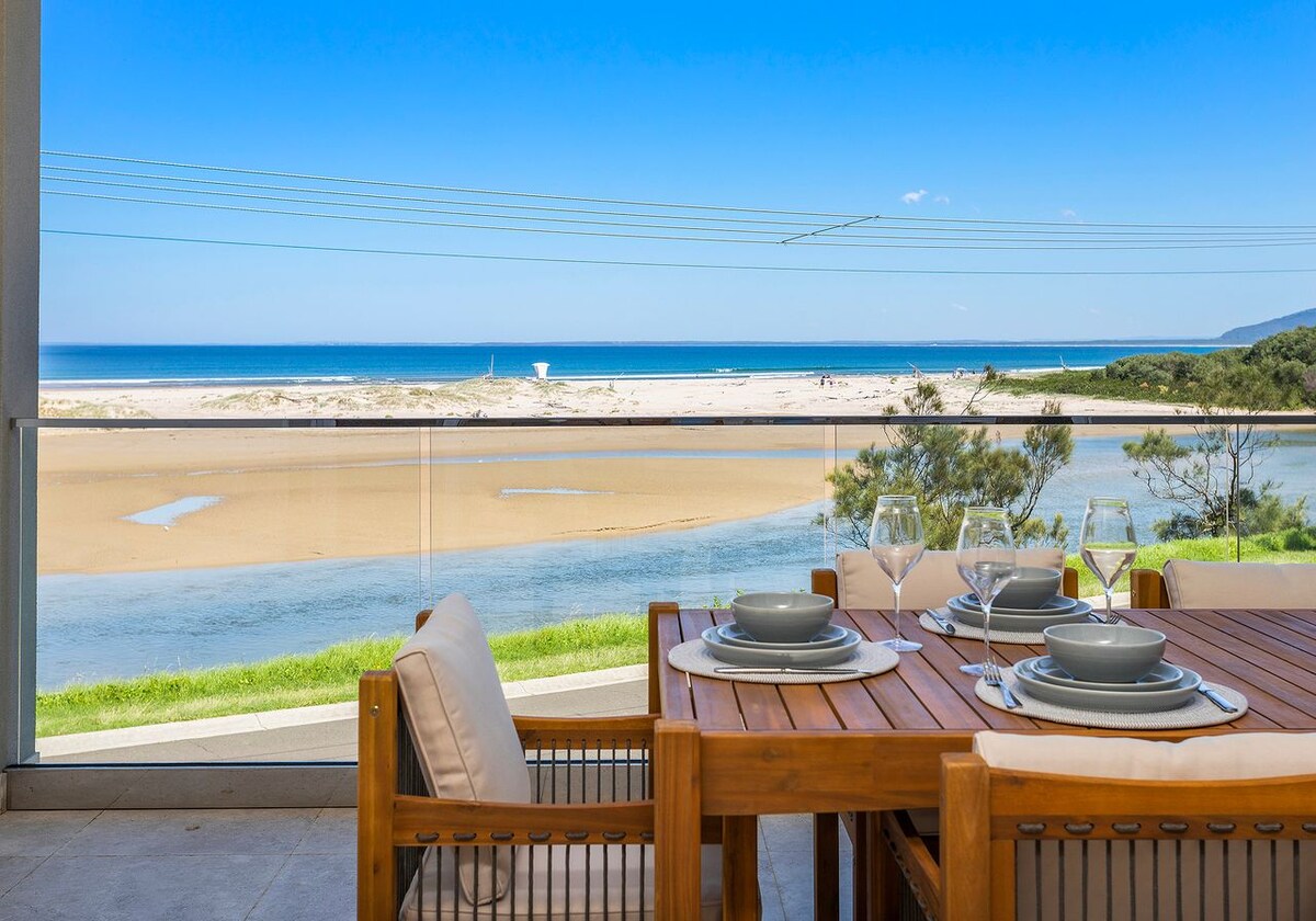 A covered outdoor dining area is shown, featuring a wooden table set for several guests. Views of the ocean and Crooked River are visible in the background, with sandy beach stretching along the shore. Blue skies complement the serene coastal landscape.