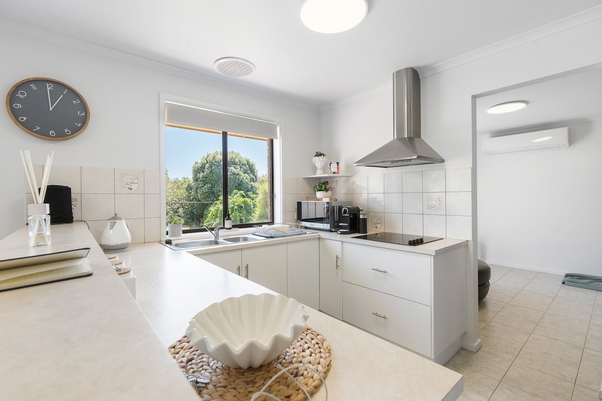 A modern kitchen is presented, featuring white cabinetry and a sleek countertop. Essential appliances, including a coffee maker and an electric cooktop, are visible. A large window provides natural light and a view of greenery, enhancing the spacious and functional layout.