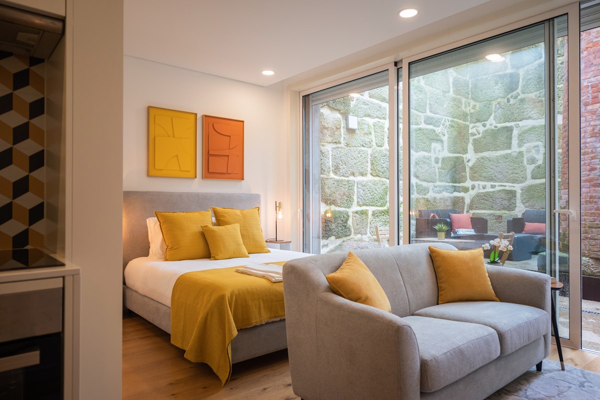 A well-lit bedroom features a spacious bed dressed in bright linens, complemented by two abstract framed artworks on the wall. A modern gray sofa with yellow accents is positioned nearby, and large glass doors offer views of a stone wall and a private outdoor seating area.