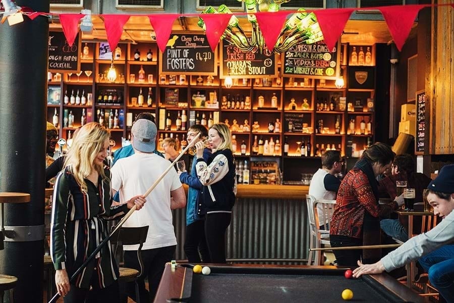 A lively bar scene showcases a billiard table in the foreground, with colorful pool balls arranged on the felt surface. A bustling backdrop includes shelves lined with various liquor bottles, illuminated by soft lighting. Red bunting adds a festive touch, enhancing the welcoming atmosphere.