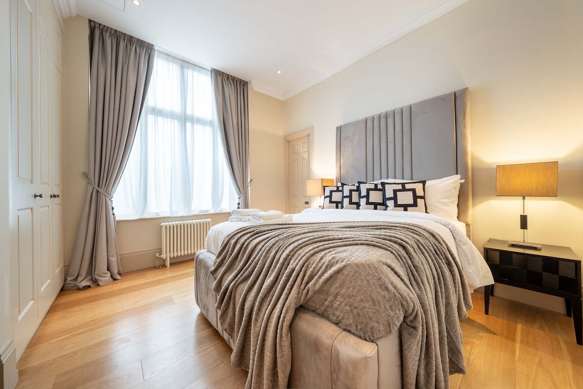 A spacious bedroom features a large bed with a light grey headboard and white linens. Soft grey curtains frame the tall window, allowing natural light to illuminate the room. A cozy throw blanket is draped over the bed, complemented by bedside tables with lamps.