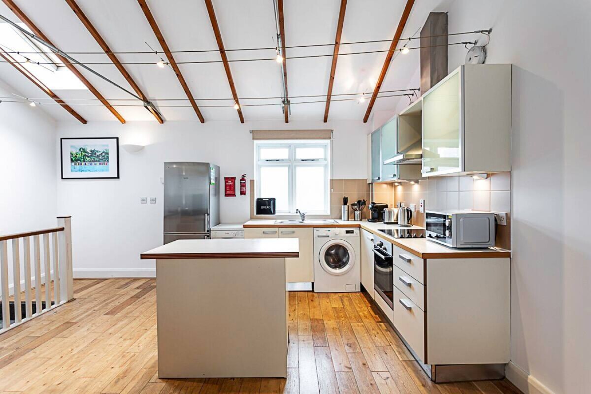 The kitchen space features modern cabinetry and stainless steel appliances, including a refrigerator, microwave, and washing machine. A central island offers additional counter space. Natural light filters in through the window, illuminating the wooden flooring and exposed beam ceiling.