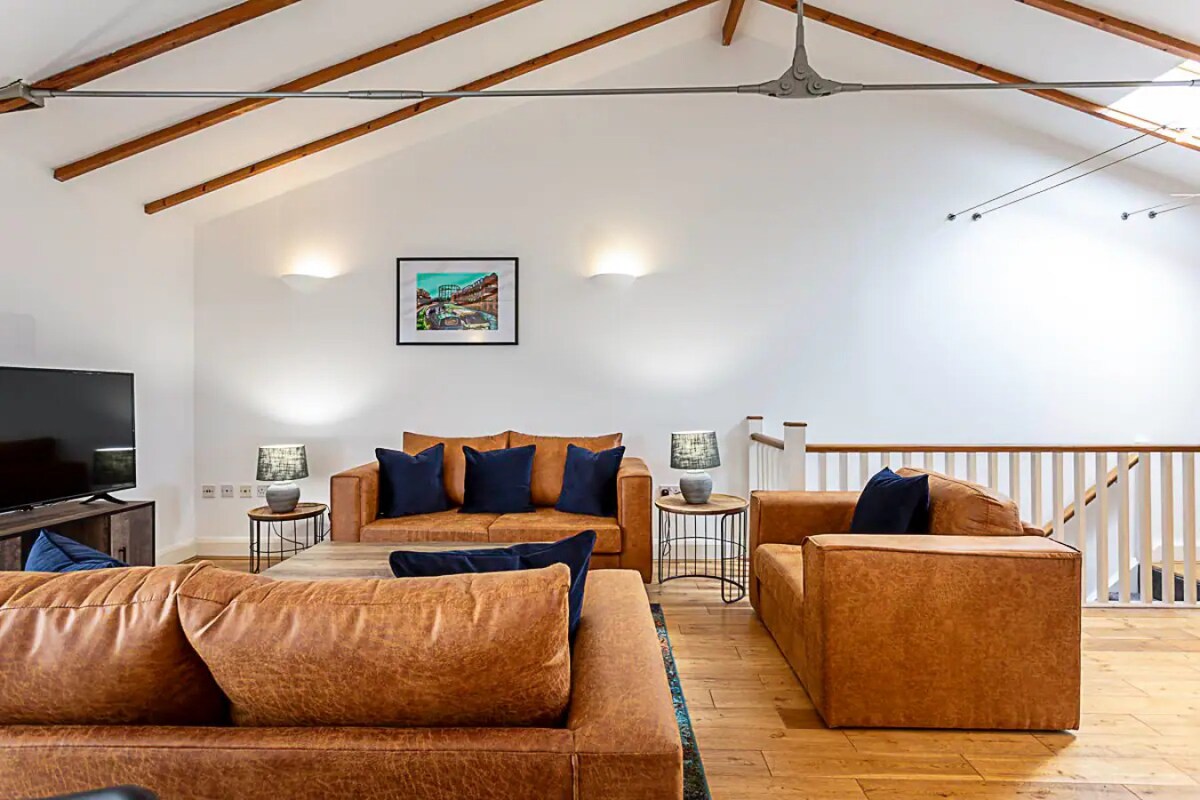 The living room features a spacious seating area with leather sofas and armchairs, complemented by soft cushions. A flat-screen TV is positioned across from the seating. High ceilings with wooden beams contribute to an airy atmosphere, while artwork adorns the wall.