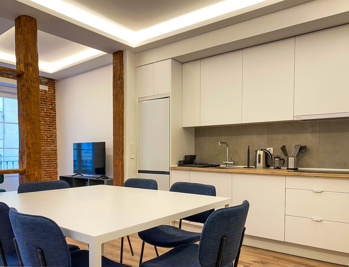 A modern kitchen and dining area is displayed, featuring a white table surrounded by six soft blue chairs. Sleek cabinetry and a fully equipped countertop accommodate various kitchen appliances, including a kettle and coffee maker. Natural light filters through a nearby window, highlighting exposed brick walls.
