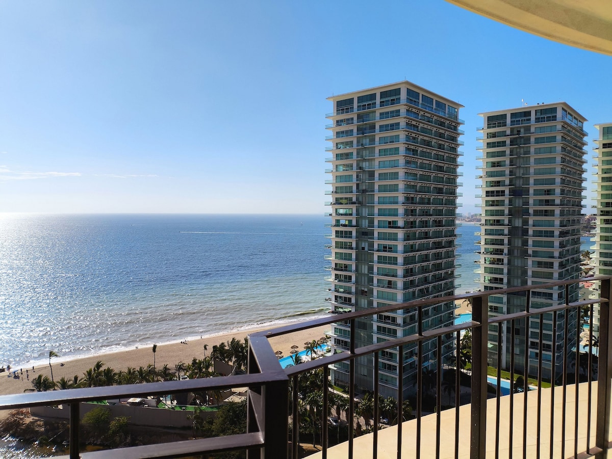 A panoramic view of the ocean is captured from a terrace, showcasing sandy beaches and glistening water. Modern beachfront buildings are visible in the foreground, with a clear blue sky above, emphasizing the tranquility of the location.
