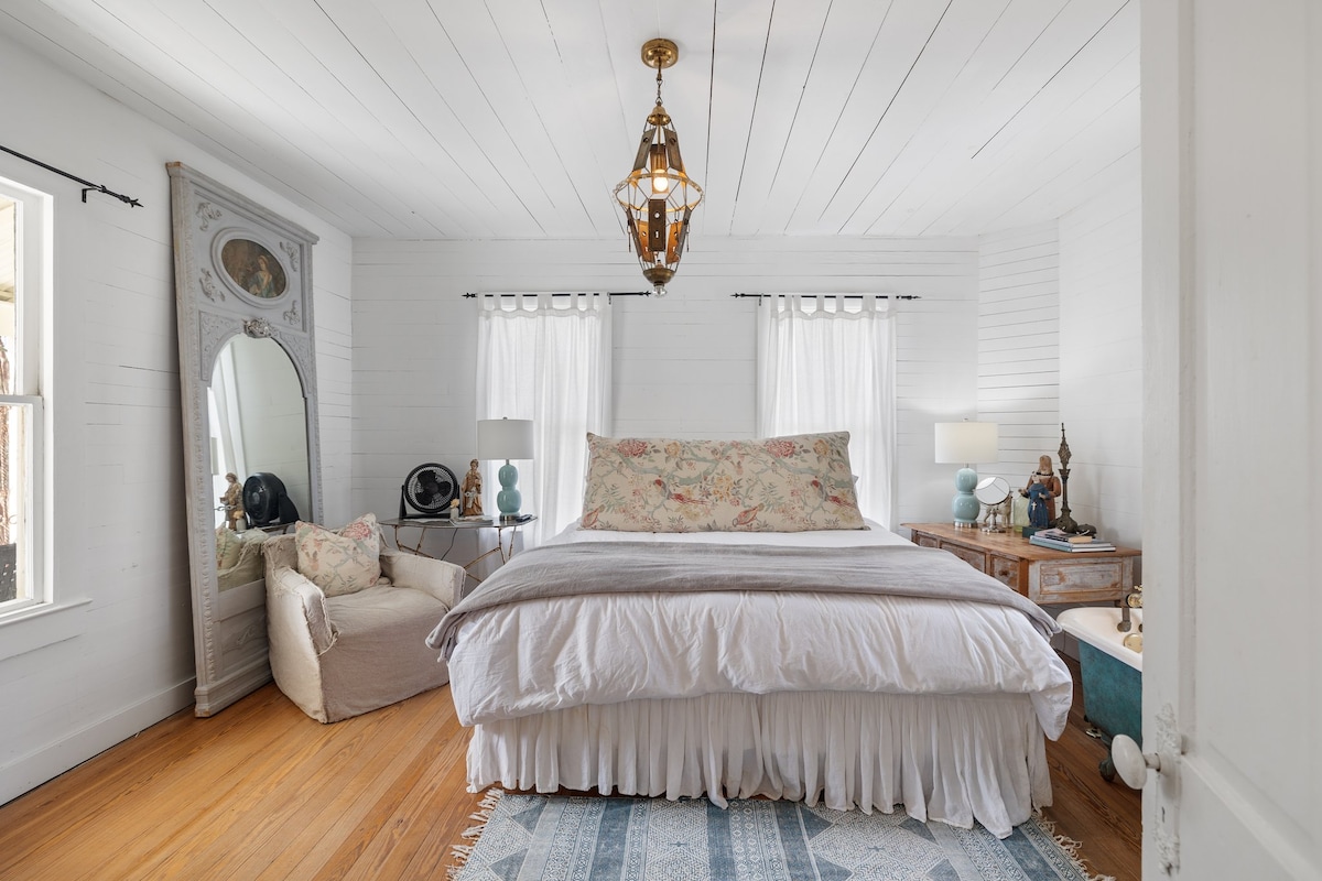 The main bedroom features a spacious bed with a light-colored bedspread and floral accents. Natural light enters through two sheer-curtained windows, illuminating a corner with a cozy armchair and a decorative mirror. Two bedside lamps provide soft lighting on either side of the bed.