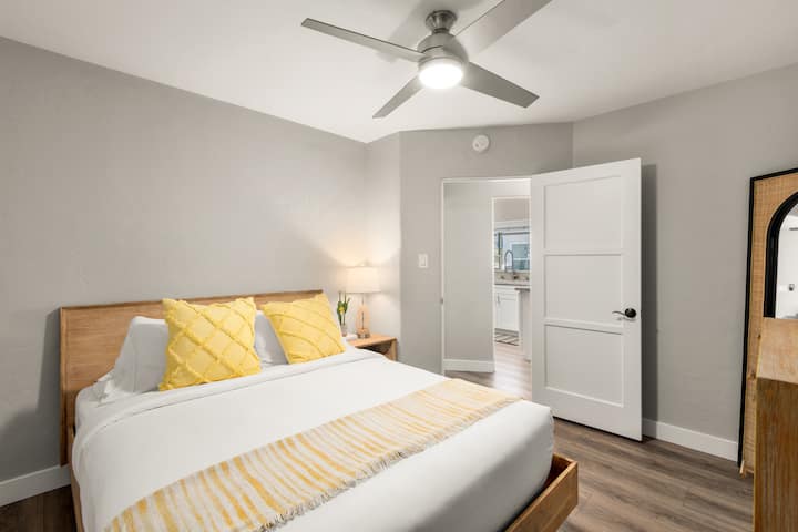 غرفة نوم مريحة مع إطار سرير خشبي وخزانة ملابس متطابقة وتلفزيون مثبت. تضيء اللمسات الصفراء الناعمة الألوان المحايدة. توفر مروحة السقف تدفق الهواء، وتوفر مصاريع المزارع التحكم في الضوء.