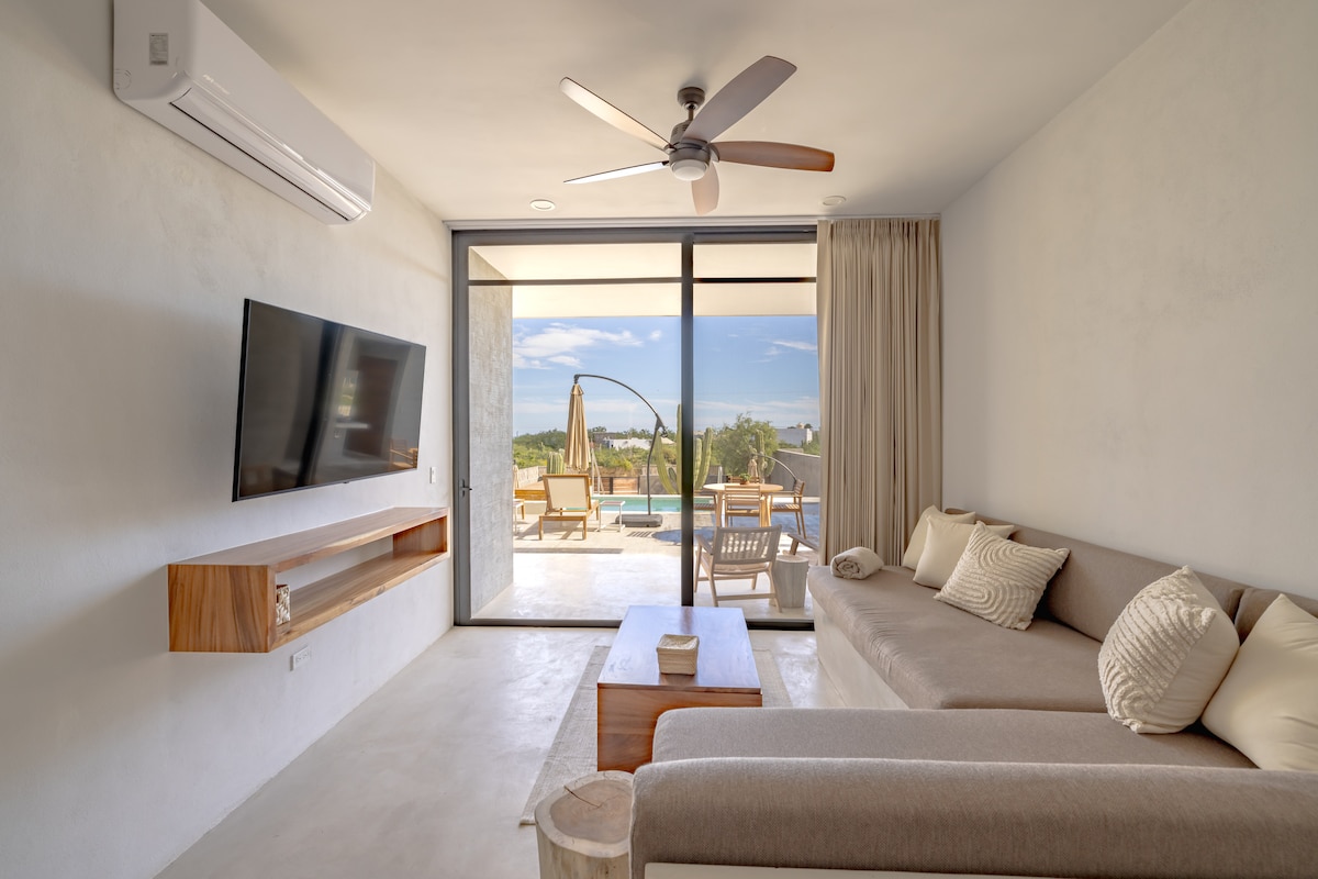 A modern living room features a spacious sofa with neutral upholstery, a flat-screen TV mounted on the wall, and a wooden shelf beneath it. Large glass doors open to a view of the pool area, allowing natural light to fill the space.