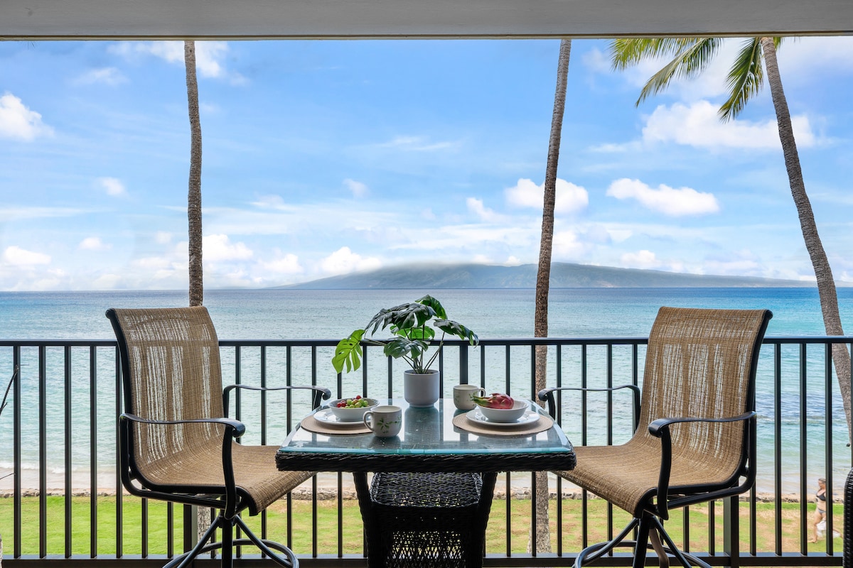 A furnished lanai features a high-top dining table set for two, overlooking the ocean. Lush palm trees frame the panoramic views of the gently lapping waves against the shore, with the distant island of Lanai visible in the background under a clear blue sky.