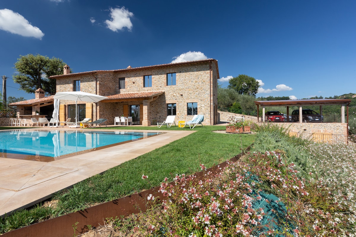 A tranquil setting features a spacious stone villa beside an inviting infinity pool, surrounded by well-maintained gardens. The property boasts a shaded outdoor area with sun loungers and a carport, offering a picturesque view of the lush landscape and blue sky.