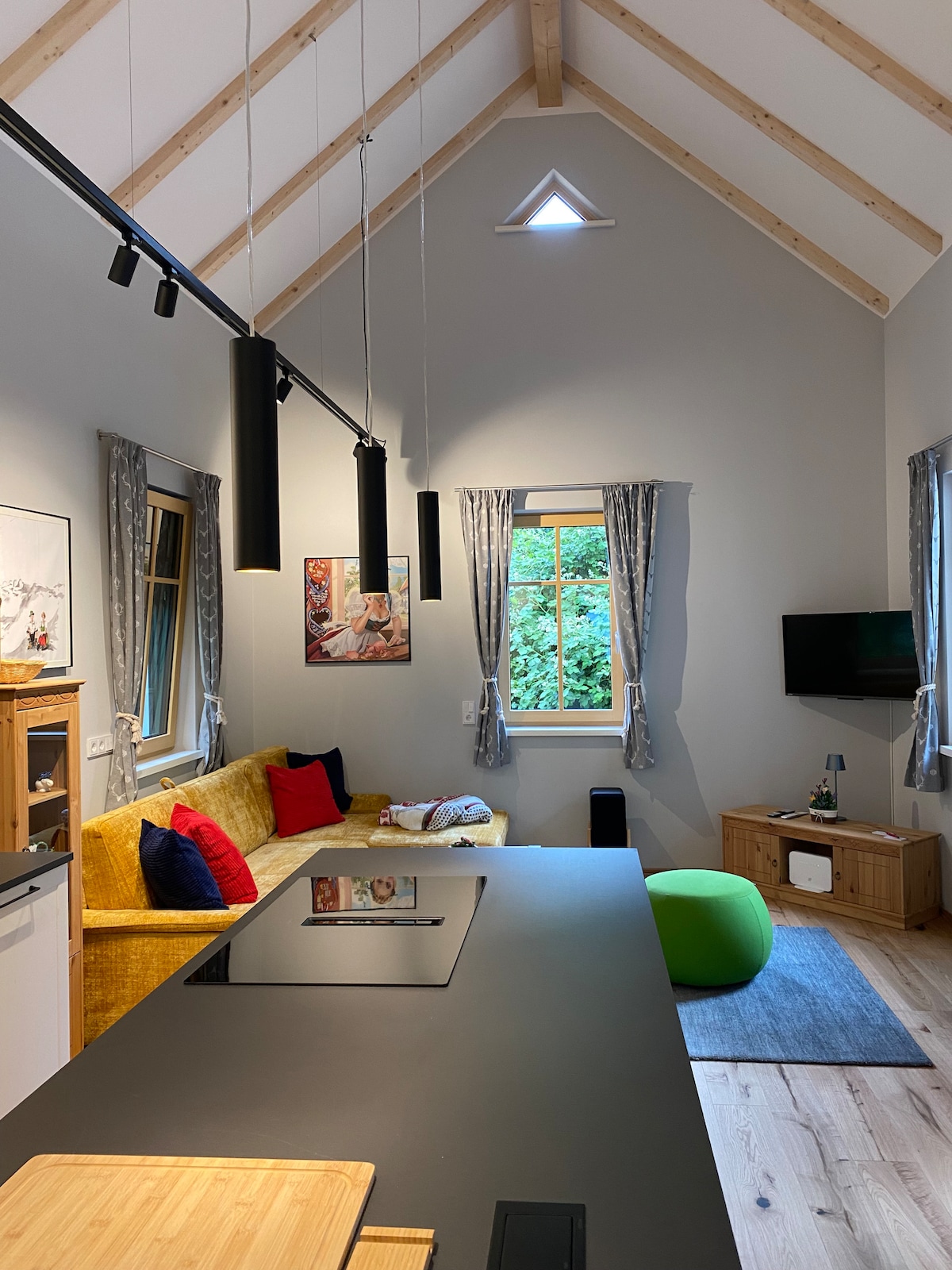 A modern living space features a high ceiling with exposed wooden beams. A cozy yellow couch is complemented by colorful cushions, while a large window allows natural light to fill the room. An entertainment area includes a flat-screen TV and a small wooden cabinet.