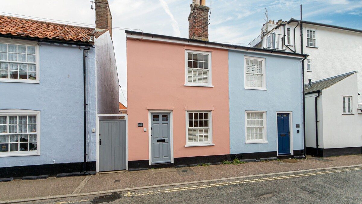 A charming cottage exterior is displayed, featuring a combination of pastel pink and blue walls. The front door is painted grey, and windows are adorned with white frames. A slate-colored door on the right complements the overall aesthetic. The setting is along a quiet street with a clear sky overhead.