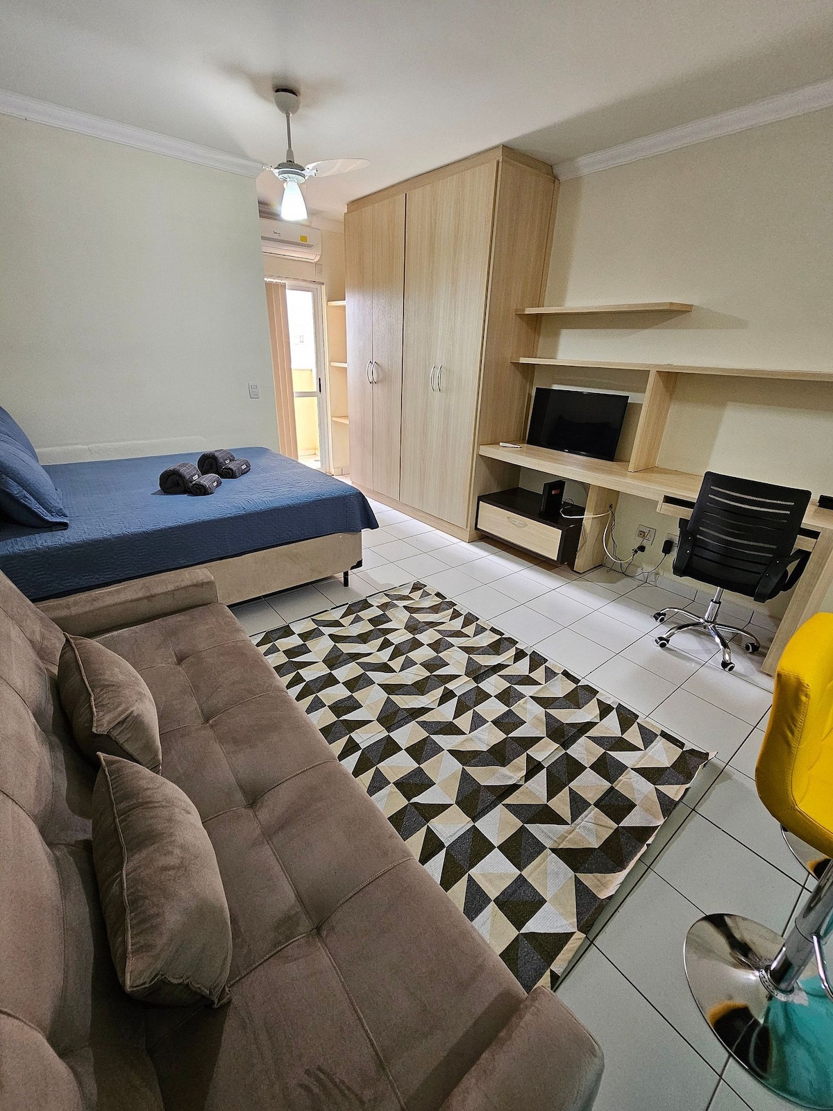 A studio interior is presented, featuring a spacious bed covered in blue bedding. A neatly arranged sofa and a patterned area rug are visible. A desk and chair are positioned next to a television unit, with a wardrobe integrated into the light-colored wooden paneling.