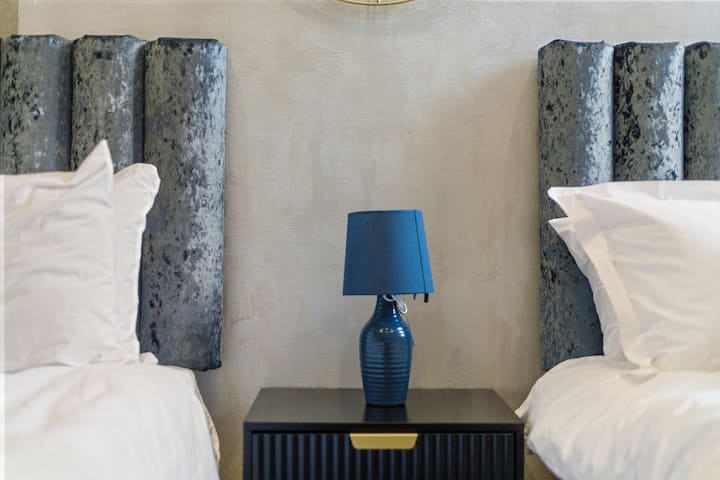- Elegante mesita de noche con lámpara moderna entre dos acogedoras camas individuales.  
- Las cabeceras suaves y las sábanas limpias crean un ambiente acogedor y tranquilo.  
- Dormitorio de Airbnb diseñado para la comodidad de amigos, familiares o grupos.