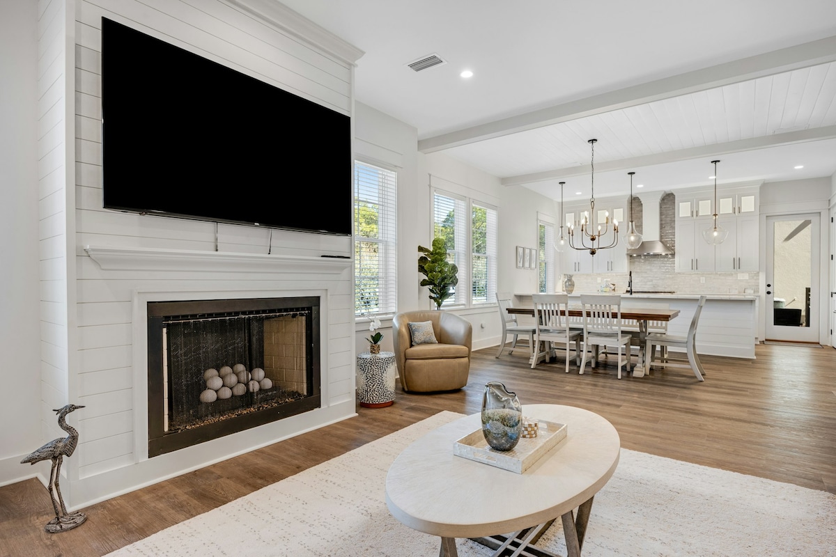 A modern living area features a spacious layout with a large wall-mounted TV above a fireplace. A cozy armchair is positioned near a light-colored coffee table. The dining area, with a long table and white chairs, is visible in the background along with a beautifully designed kitchen.